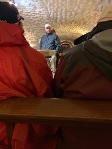 Man speaking to a group of people in a dimly lit room with a low, carved ceiling