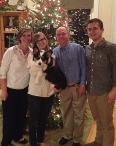 Mom, college aged daughter holding a black, white and tan corgi, dad and college aged son standing in front of a Christmas tree