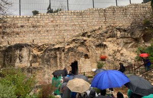People with umbrellas walking down steps towards an empty tomb in a rocky wall.